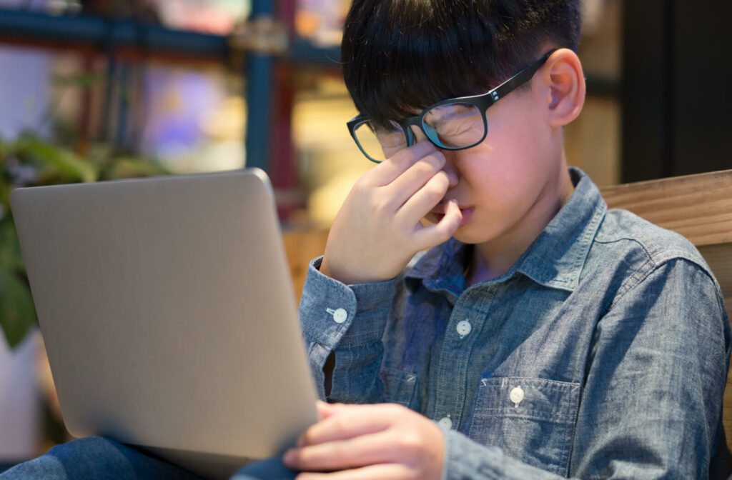 A boy wearing eyeglasses is squeezing his nose bridge as his eyes are strained and stressed due to long exposure from his computer screen.