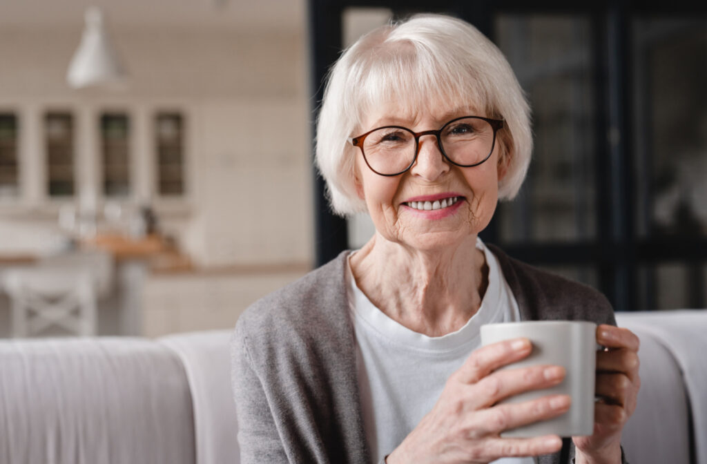 A senior woman with glasses and short white hair smiling, sitting in a couch as she looks directly at the camera