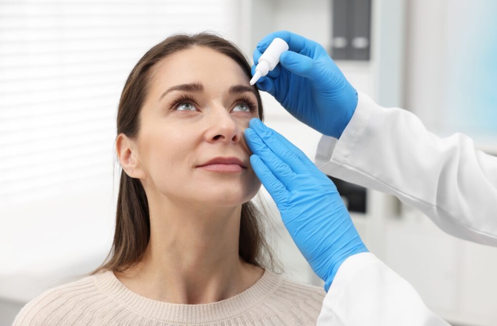 A healthcare professional wearing blue gloves administering eye drops to a woman in a clinical setting, with the patient looking upward.