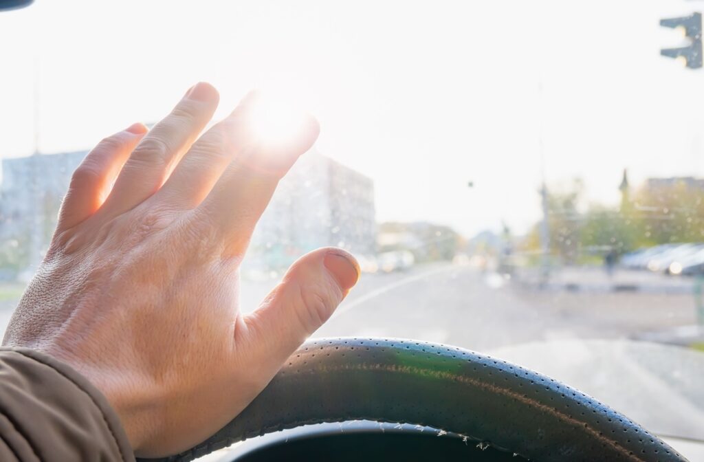 A first-person view of an adult driving a car through a city, raising a hand to block out a blinding glare in their windshield.