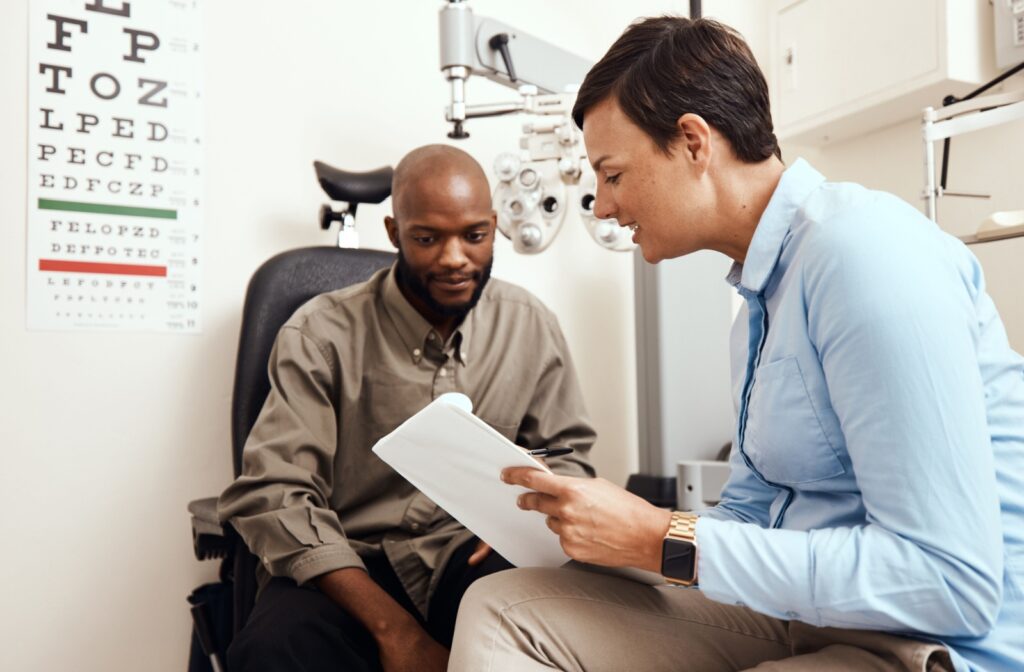 An optometrist holding a clipboard talking to a patient after an eye exam and discussing what's causing them to see a glare.