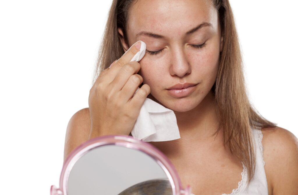 A person looking in a table top mirror, using a cleansing wipe to clean their eyelid as part of a healthy eyelid hygiene routine.