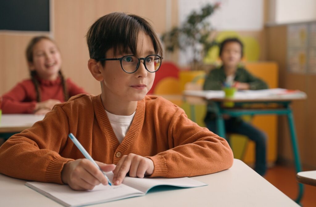 A child in a classroom sitting at a desk, wearing glasses to view the board.