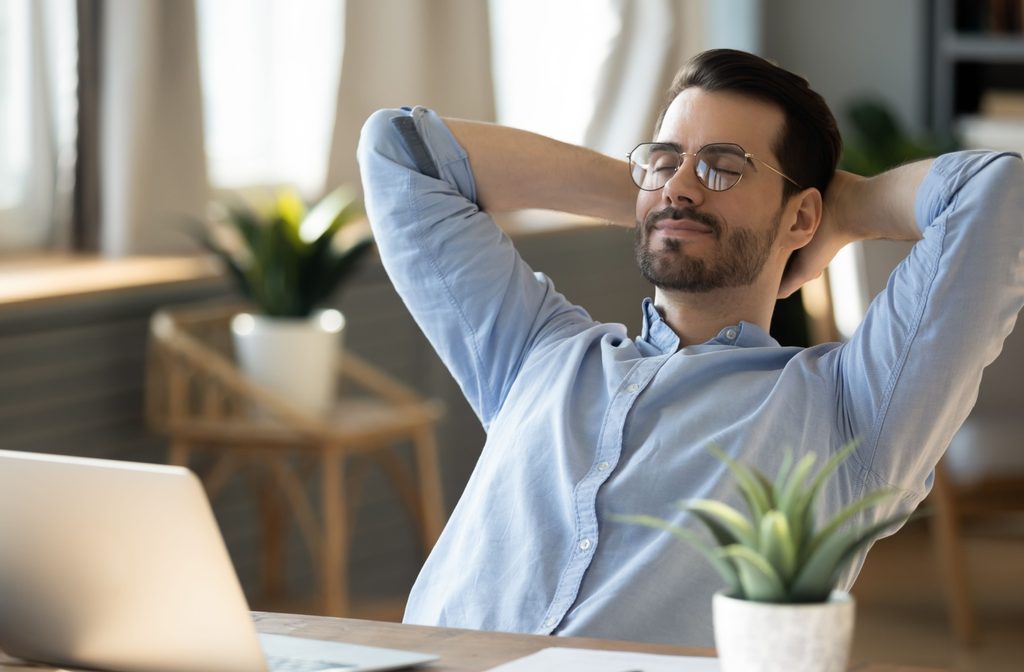 A person sitting at a desk with a laptop in front of them, leaning back with their eyes closed to give their eyes a break from the screen.